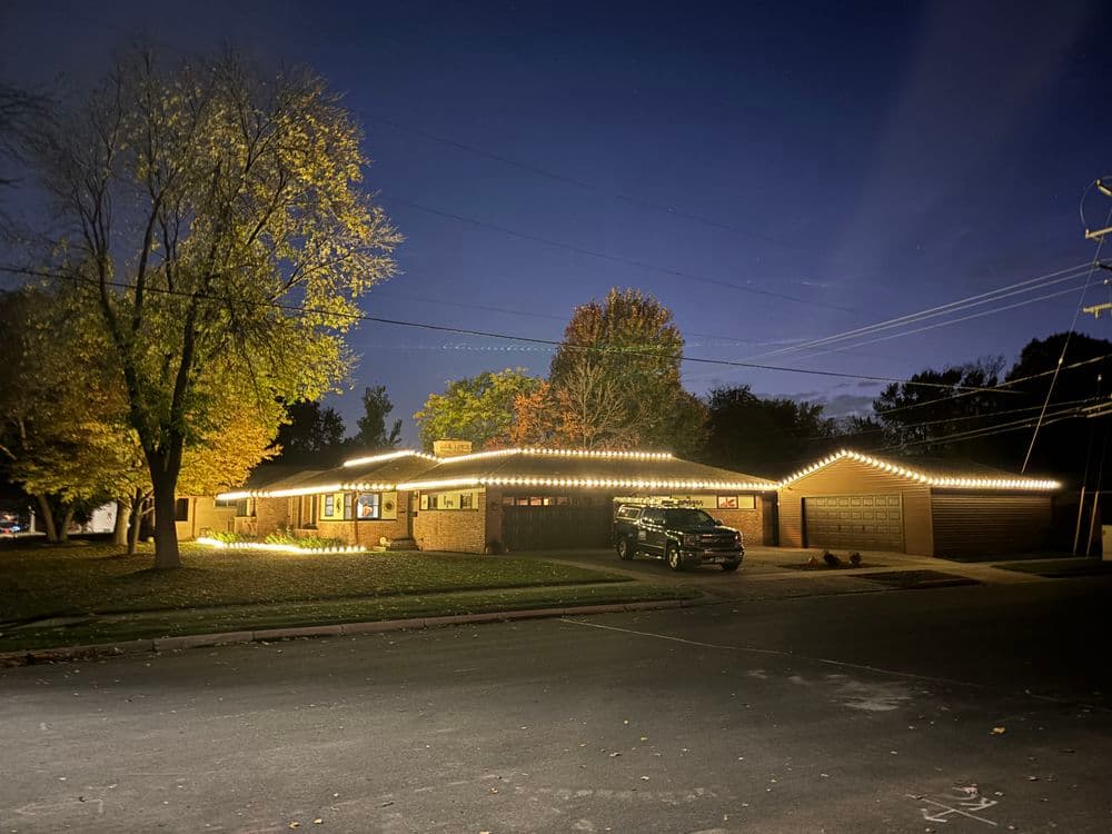 Home decorated with outdoor lights at night, surrounded by autumn trees and a street view.