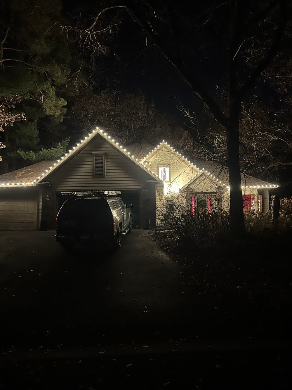Cozy house with festive holiday lights and a parked SUV in a dark, wooded setting.